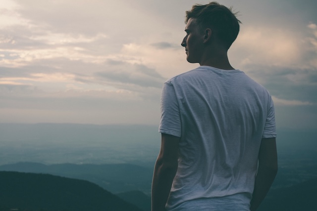 A young man standing on top of a mountain looking at the cloudy sky