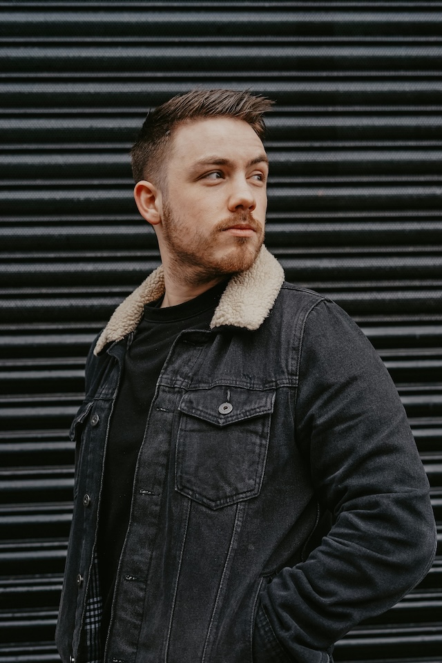 Young man standing against black garage door with his hands in his black jean jacket pockets